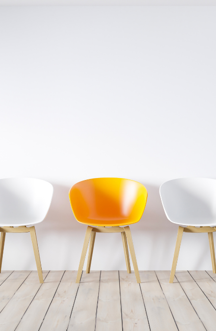 A single orange chair inbetween a row of white chairs in a white room with a white wooden floor