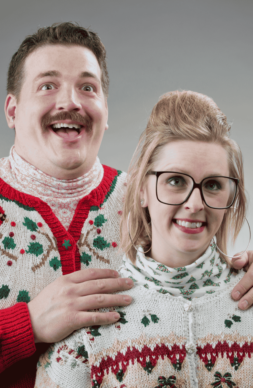 A man & a woman in 80s Christmas jumpers looking happy and nerdy